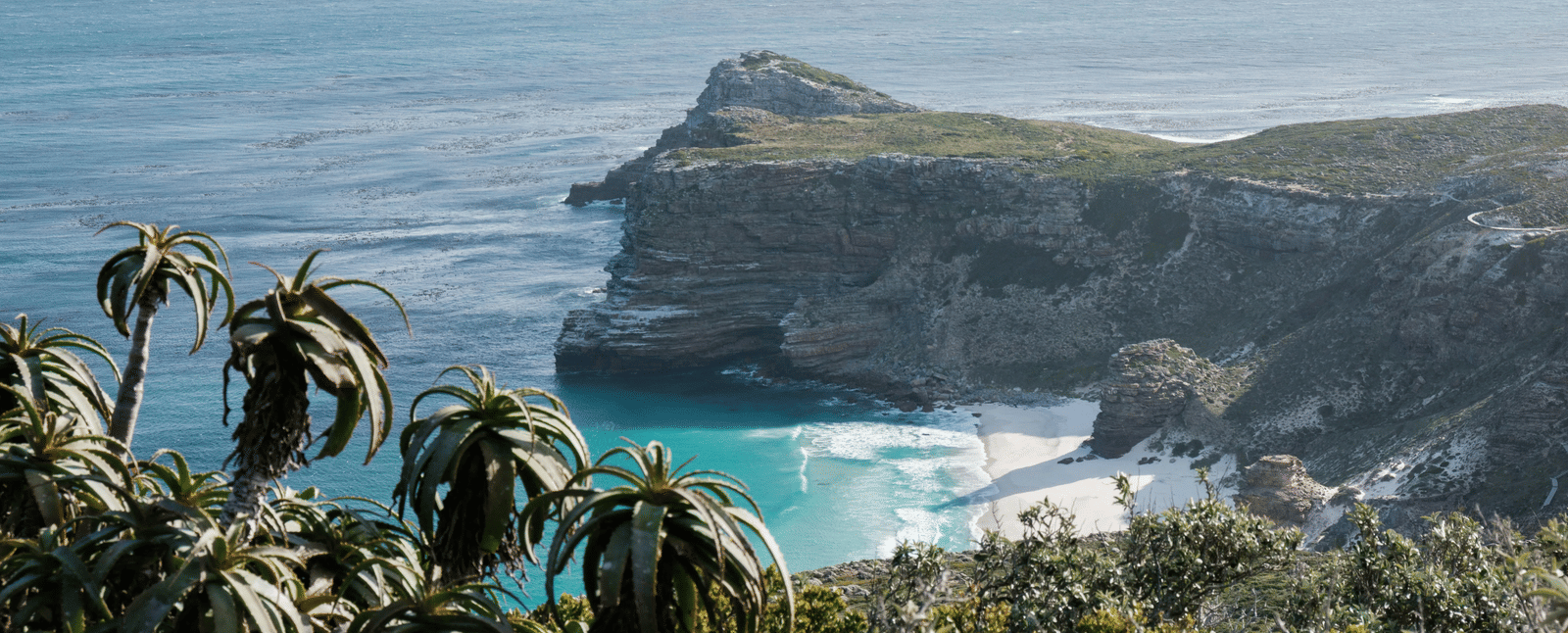 A rock cliff viewed from a far overlooking the vast turquoise ocean in front of it stretching till the end of the horizon.