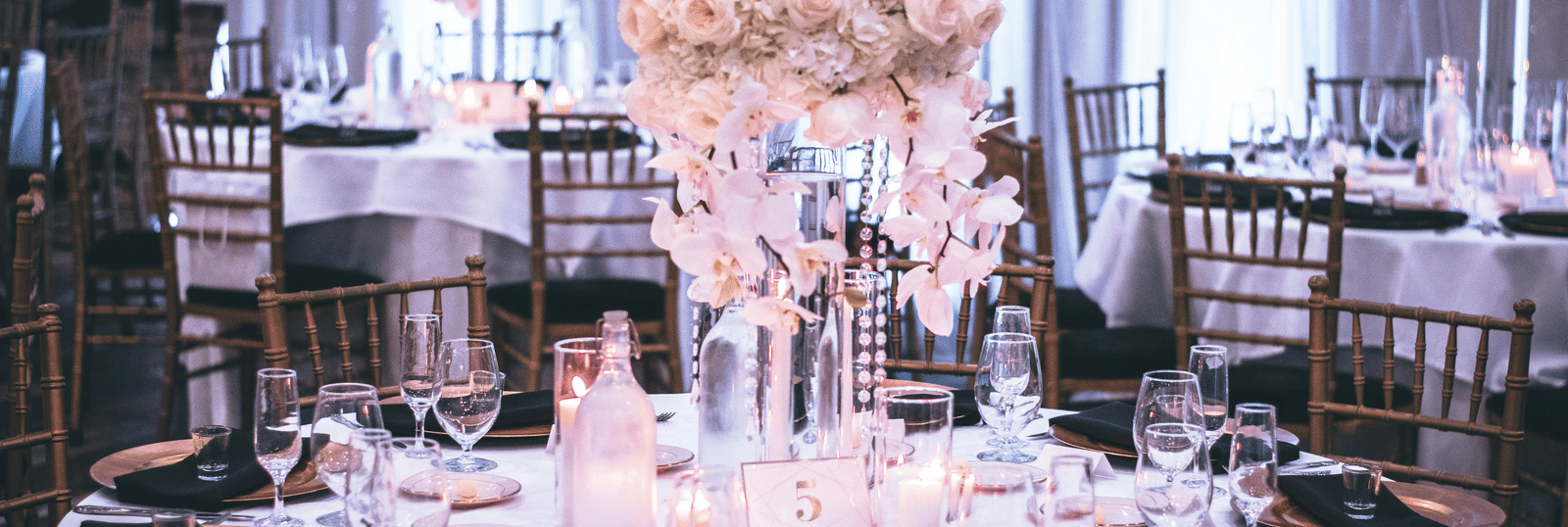 A round table with a white table cloth along with huge plates set on the table and a beautiful flower vase in the middle.