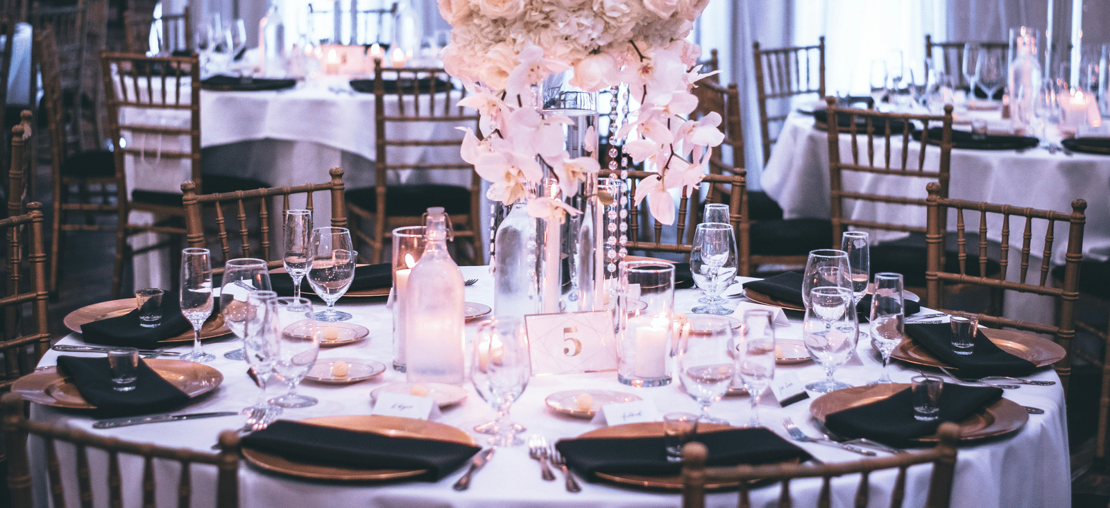 A round table with a white table cloth along with huge plates set on the table and a beautiful flower vase in the middle.