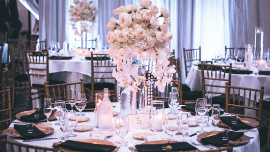 A round table with a white table cloth along with huge plates set on the table and a beautiful flower vase in the middle.