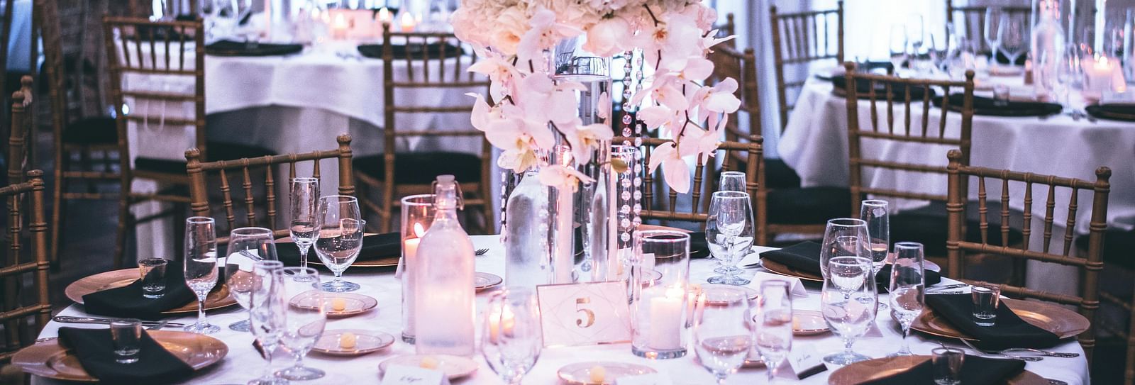 A round table with a white table cloth along with huge plates set on the table and a beautiful flower vase in the middle.