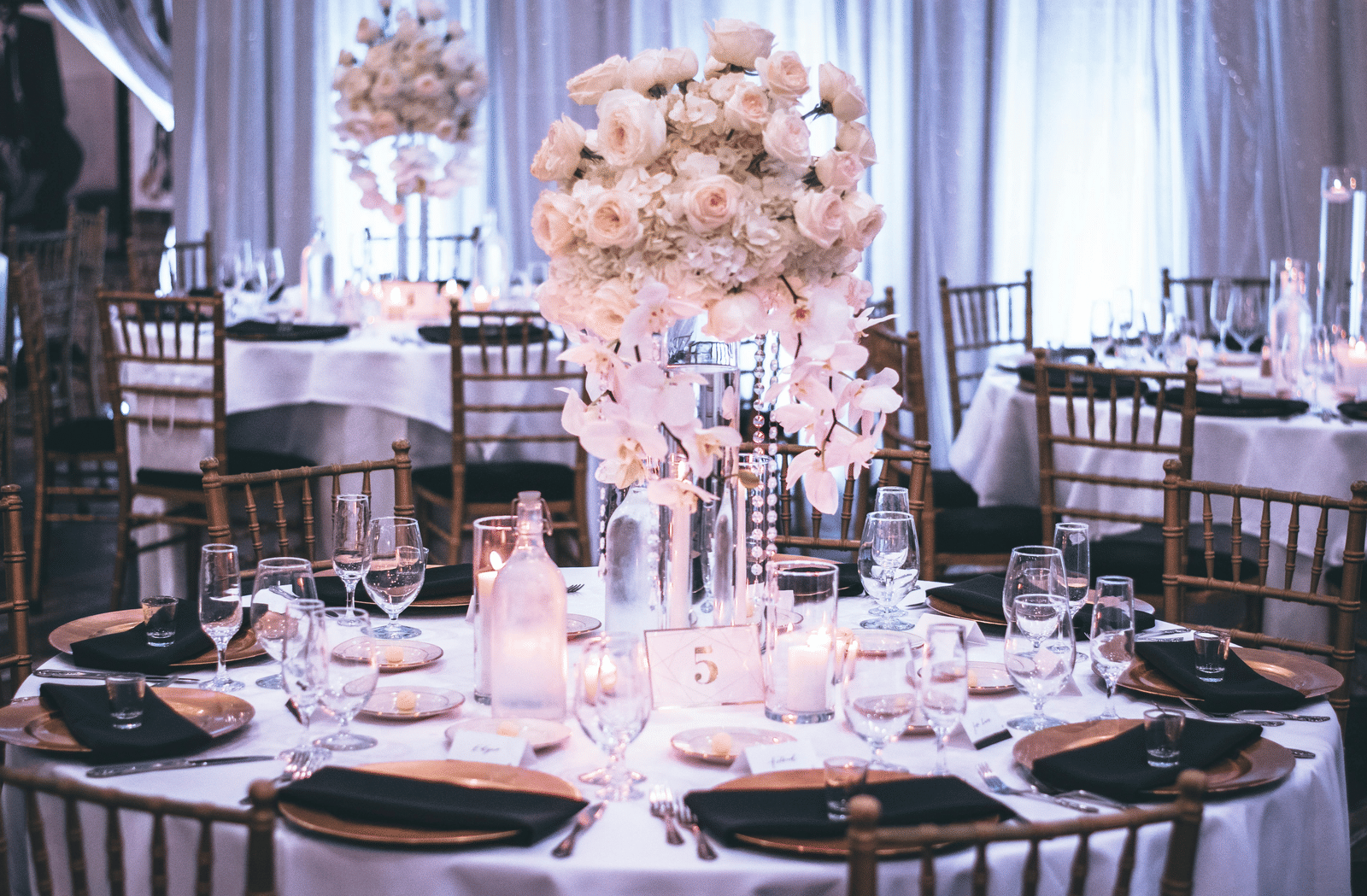 A round table with a white table cloth along with huge plates set on the table and a beautiful flower vase in the middle.