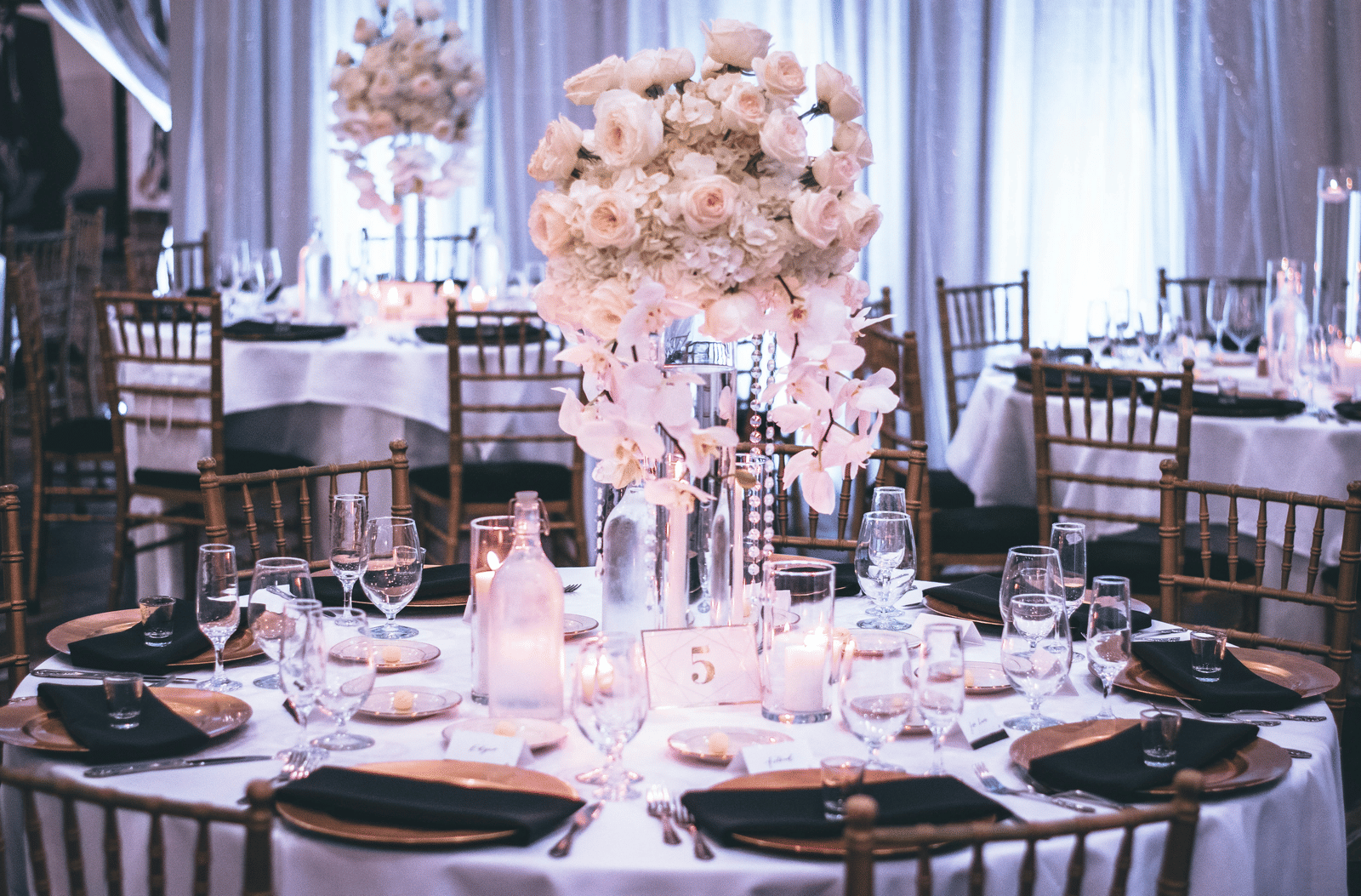 A round table with a white table cloth along with huge plates set on the table and a beautiful flower vase in the middle.