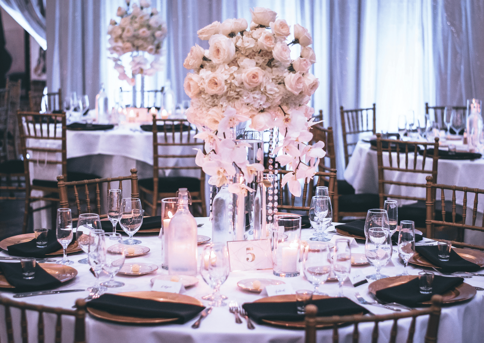 A round table with a white table cloth along with huge plates set on the table and a beautiful flower vase in the middle.