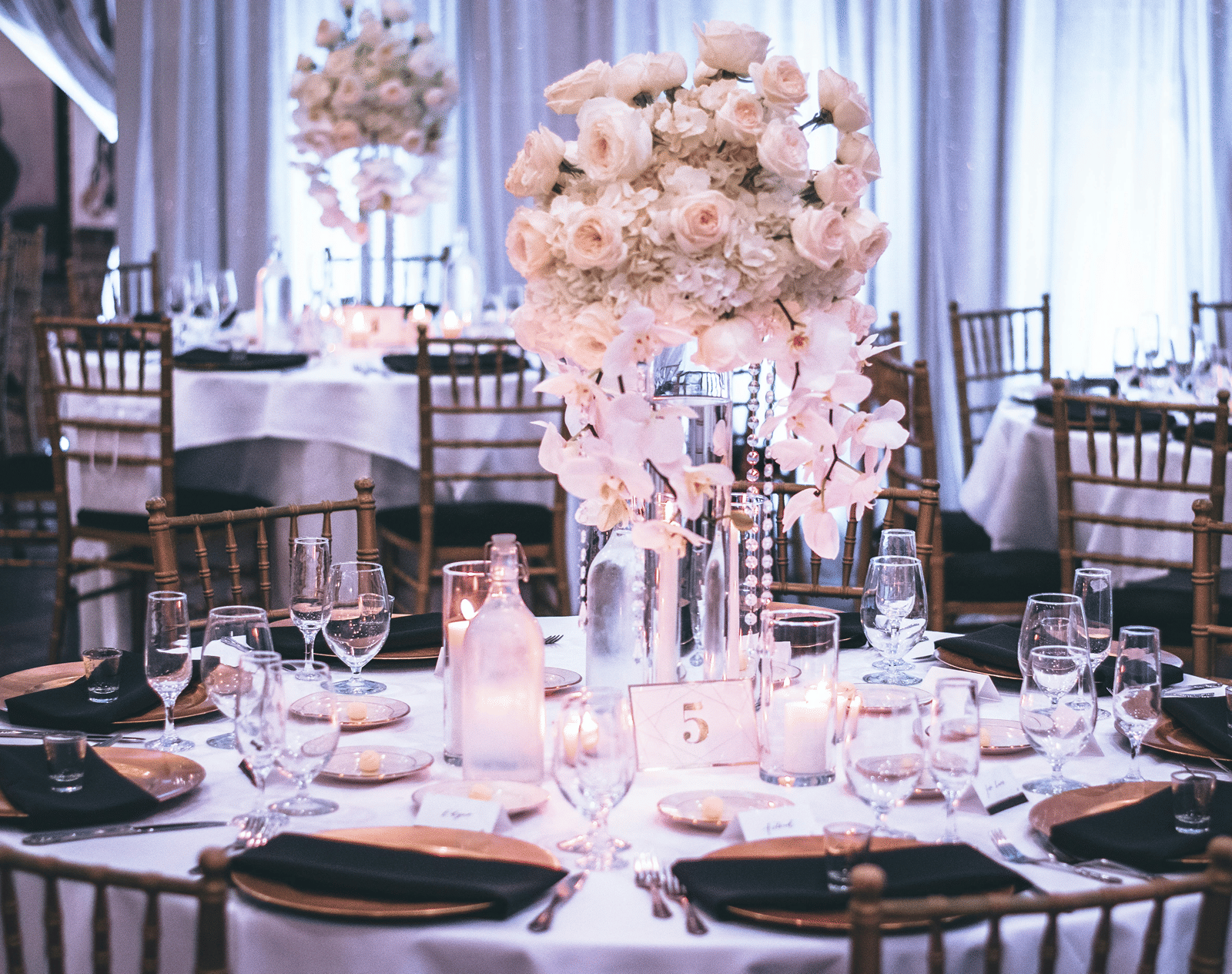 A round table with a white table cloth along with huge plates set on the table and a beautiful flower vase in the middle.