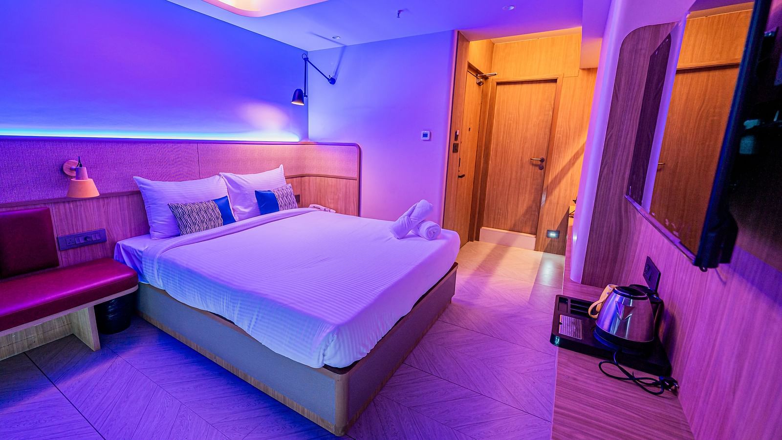 A cosy hotel room with modern lighting, a double bed, bedside lamp, wall-mounted TV, tea/coffee maker, and warm ambient decor at Indie Stays Mumbai International Airport