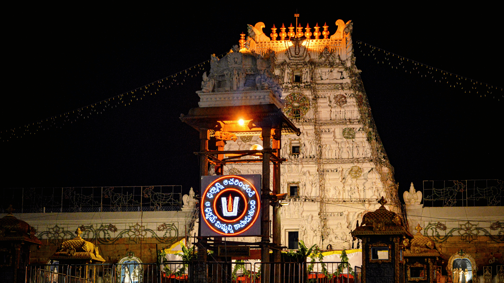 The illuminated Tirupati temple gopuram glowing against the night sky with decorative lights and temple structures visible in the foreground.