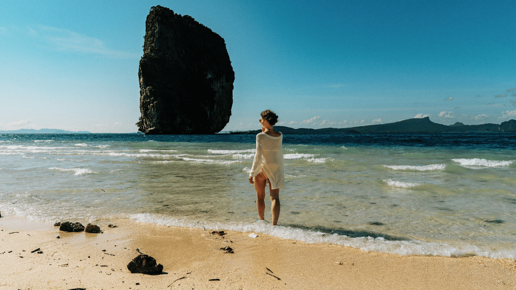 A person stands on a sandy beach looking out at a large rocky island formation in the sea.