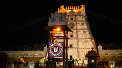 The illuminated Tirupati temple gopuram glowing against the night sky with decorative lights and temple structures visible in the foreground.