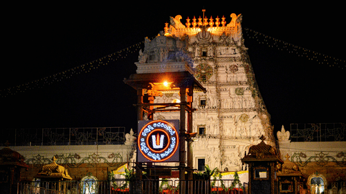 The illuminated Tirupati temple gopuram glowing against the night sky with decorative lights and temple structures visible in the foreground.