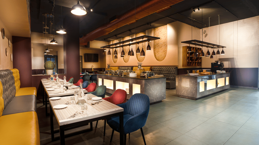 A stylish modern restaurant with colourful velvet chairs, long dining tables, and ambient hanging lights over a central service bar at Apartel by Aarin, Oragadam.