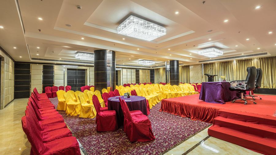 Large conference hall featuring rows of yellow chairs, a red-carpeted stage with purple tables, and elegant chandeliers at Golden Tulip Kukas, Jaipur.