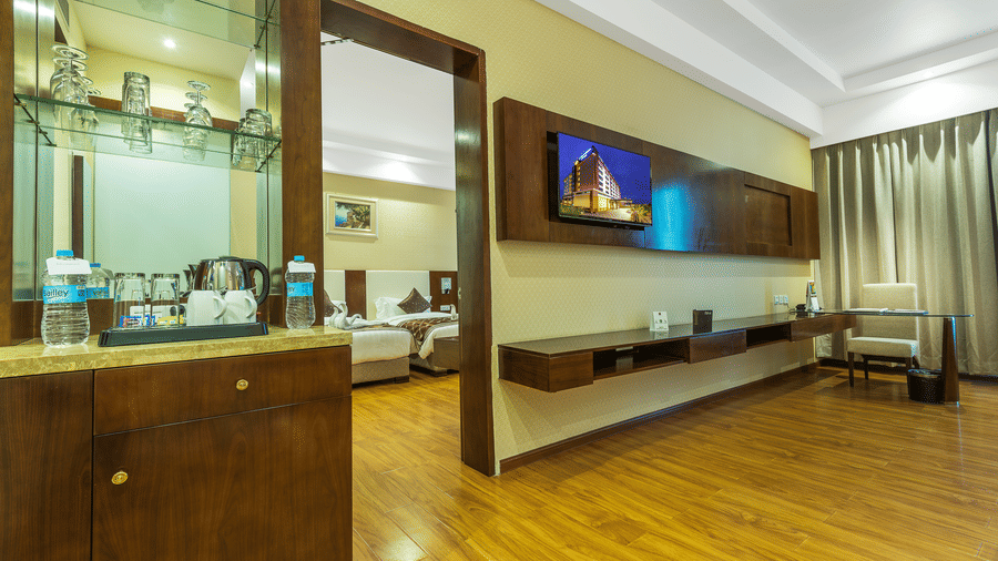 Hotel suite interior showing a wooden refreshment station and a view through a doorway into a bedroom with twin beds and a wall-mounted TV at Golden Tulip Kukas, Jaipur.
