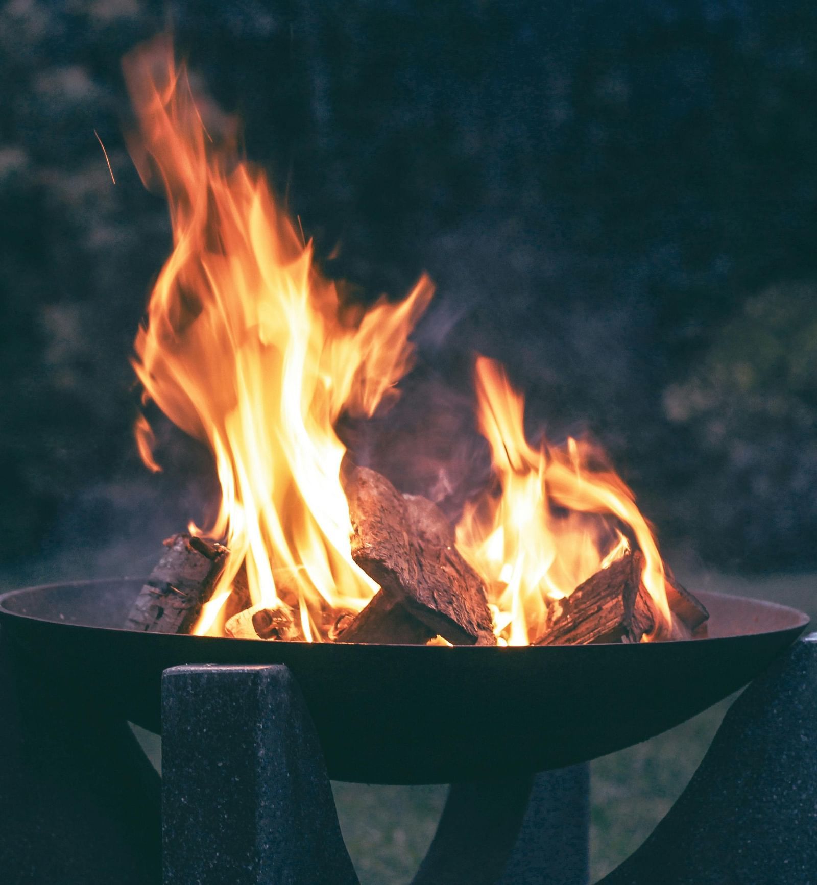 A metal fire pit with a strong flame burning inside, against a dark background of trees