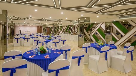 A banquet hall set up with round tables and white chairs adorned with blue sashes, featuring mirrored walls and ceiling lighting at Lords Eco Inn Morbi.