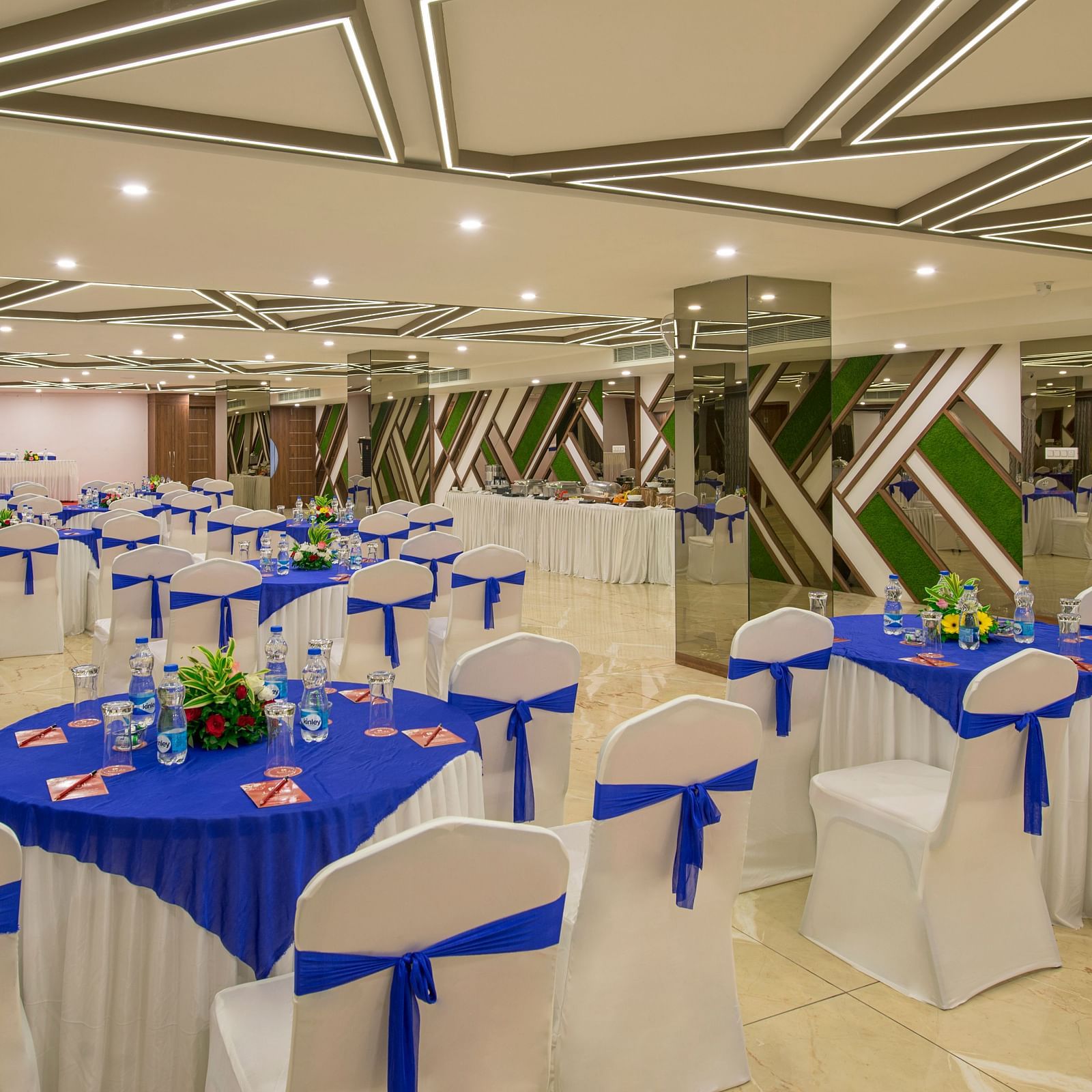 A banquet hall set up with round tables and white chairs adorned with blue sashes, featuring mirrored walls and ceiling lighting at Lords Eco Inn Morbi.
