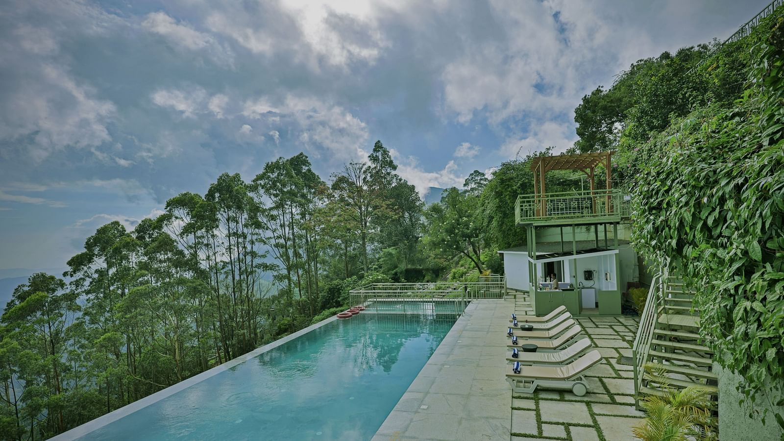 An infinity swimming pool set in the middle of a lush green area on a hillside, with sunbeds and a security watchtower nearby – Fragrant Nature Munnar