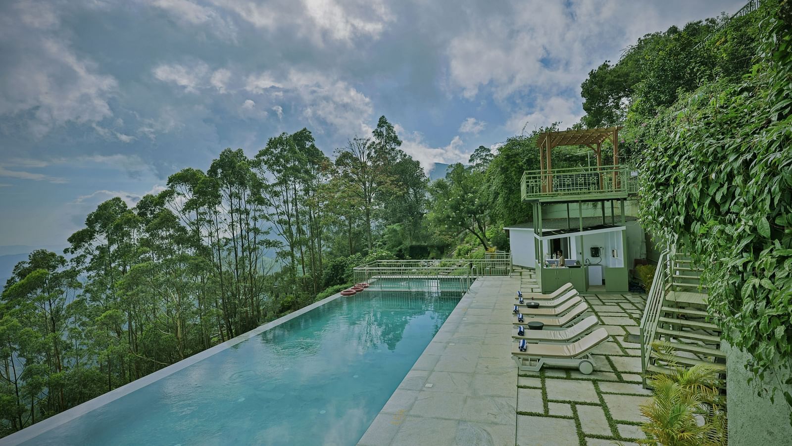 An infinity swimming pool set in the middle of a lush green area on a hillside, with sunbeds and a security watchtower nearby – Fragrant Nature Munnar