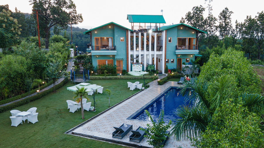 Facade of Corbett Nirvana Resort, Uttarakhand featuring two storey cottage style building, a swimming pool, and a lawn with seating arrangement.