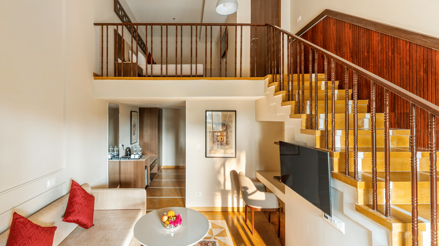 Duplex Suite at EsthereaRaj Leela, Ranakpur, featuring a seating area with red pillows, a television, and a wooden staircase leading upstairs.