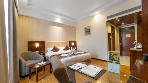 Elegant hotel bedroom featuring a double bed, a grey armchair with a coffee table, and polished wood floors under warm ambient lighting at Golden Tulip Kukas, Jaipur.
