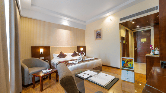 Elegant hotel bedroom featuring a double bed, a grey armchair with a coffee table, and polished wood floors under warm ambient lighting at Golden Tulip Kukas, Jaipur.