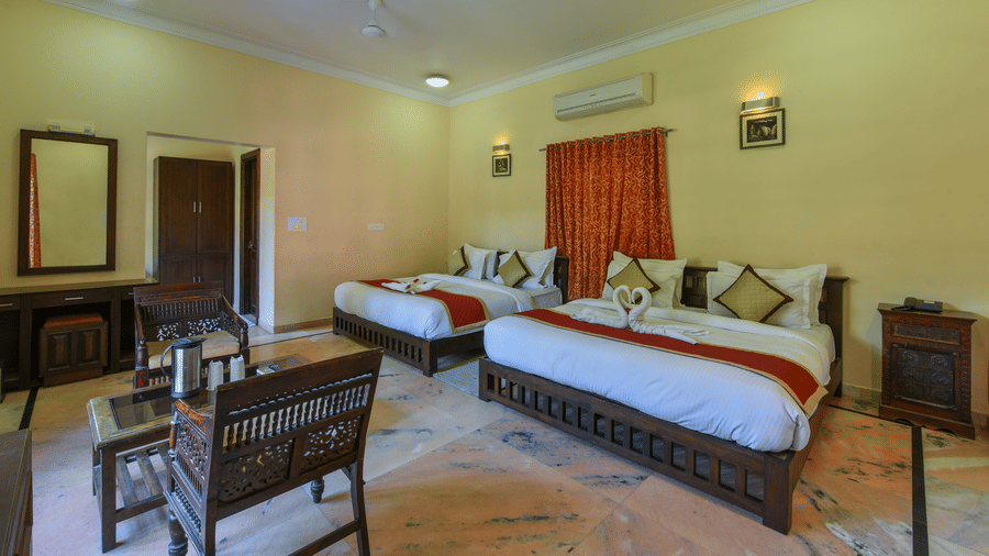 A bedroom at Garh Govind, with 2 king size beds with white linens, mirror, air-conditioning, a wooden coffee table, and armchairs.