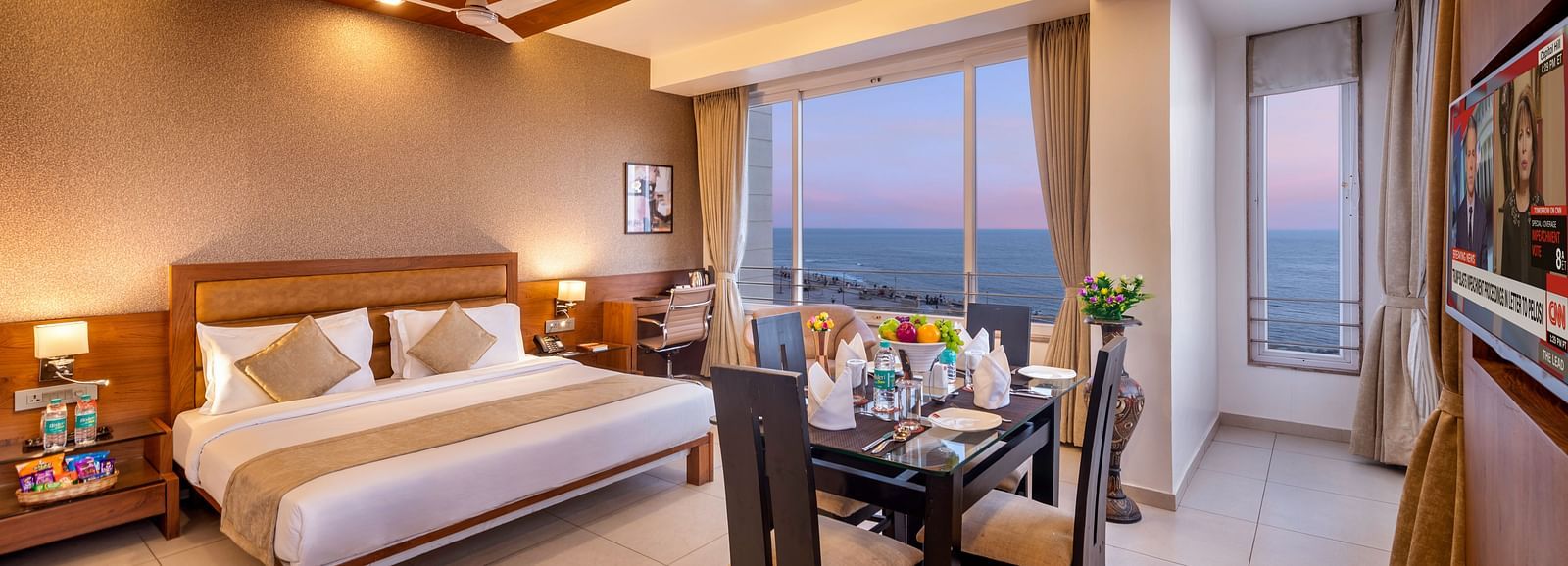 A comfortable hotel room with a bed, dining area, workspace, and a large window offering a view.