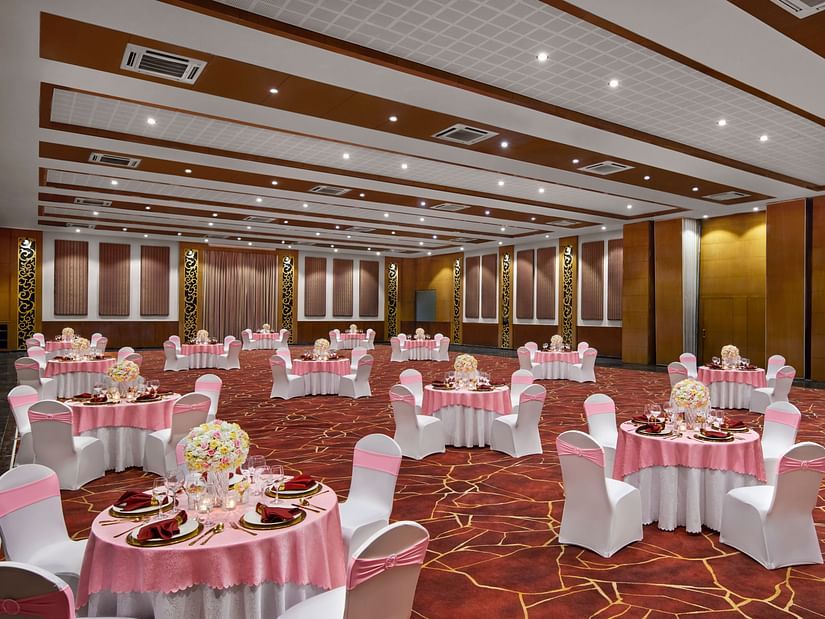 A large ballroom is elegantly set with round tables, pink tablecloths, and white chairs with pink sashes at DGV Resort by Treat, Silvassa