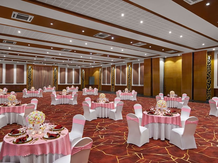 A large ballroom is elegantly set with round tables, pink tablecloths, and white chairs with pink sashes at DGV Resort by Treat, Silvassa