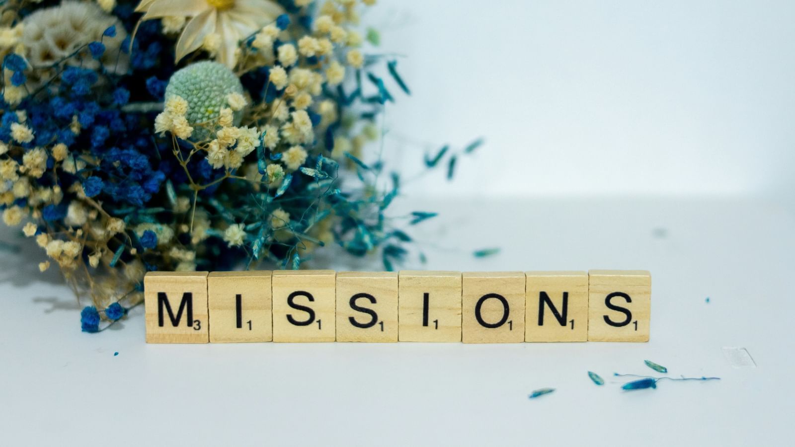 The word MISSIONS spelled out in wooden scrabble-like tiles, placed in front of a blurred background of flowers. 