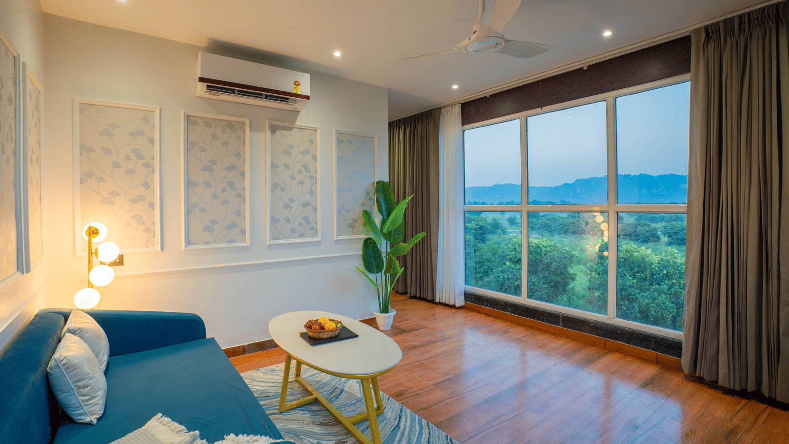 A guest room seating area at Sukoon by the Ganges with a blue sofa, modern lamp, and large window overlooking the countryside.