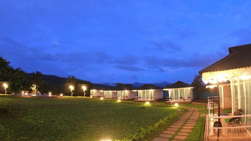 Night view of Nature Trails Durshet resort property, featuring illuminated cottages or tents, neatly bordered by pathways and expansive green lawns under a twilight sky.