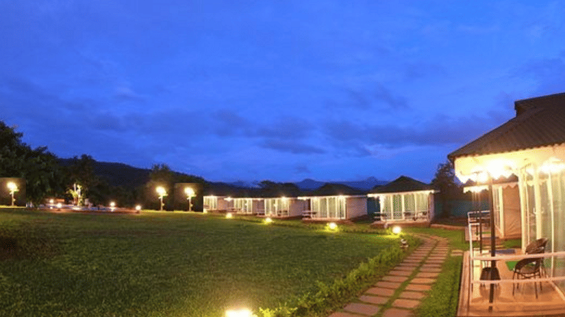 Night view of Nature Trails Durshet resort property, featuring illuminated cottages or tents, neatly bordered by pathways and expansive green lawns under a twilight sky.
