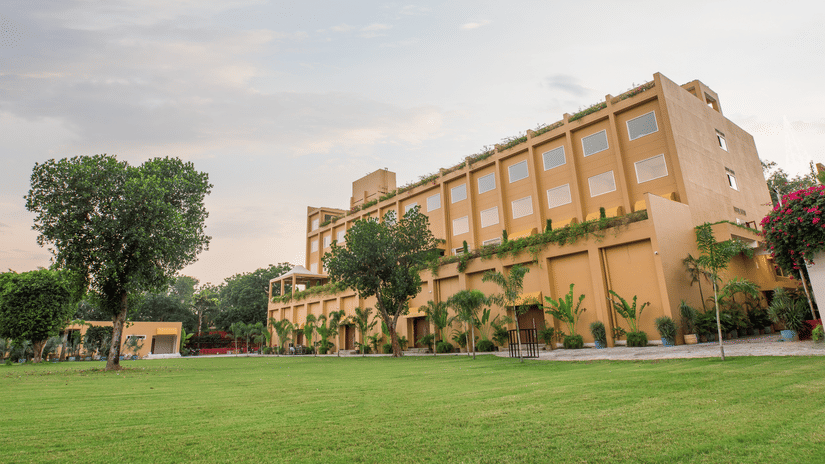 A wide-angle view of The Lalita Grand Mathura - Vrindavan, a modern multi-story hotel with a beige facade, large windows, and lush green landscaping. The foreground features a well-maintained lawn with palm trees and flowering plants, under a serene sky at