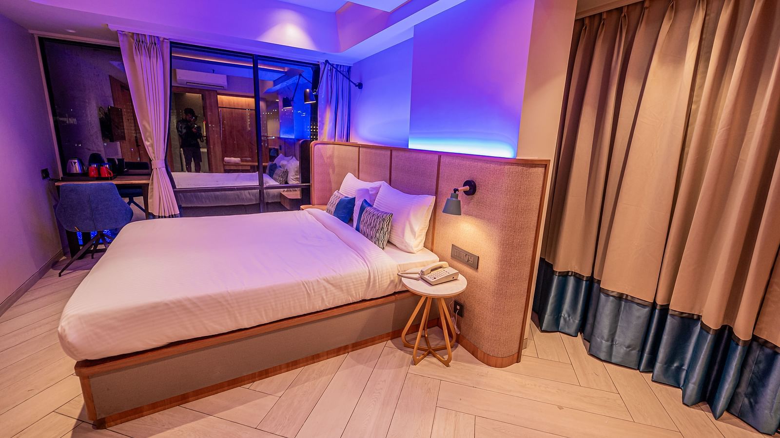 A spacious room at Indie Stays Mumbai International Airport features a large bed with a wooden headboard, a three-legged table with a lamp, and a soothing light.