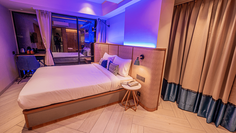 A spacious room at Indie Stays Mumbai International Airport features a large bed with a wooden headboard, a three-legged table with a lamp, and a soothing light.