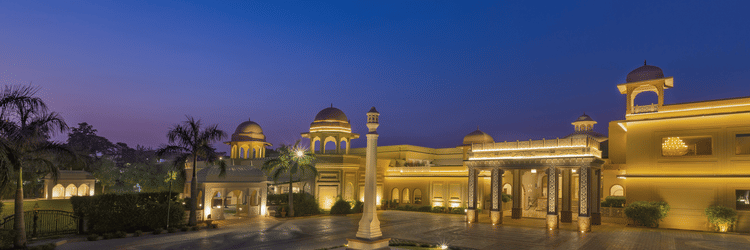 Front view of Heritage Village Resorts & Spa, Manesar with driveway, central fountain, and landscaped entrance in night.