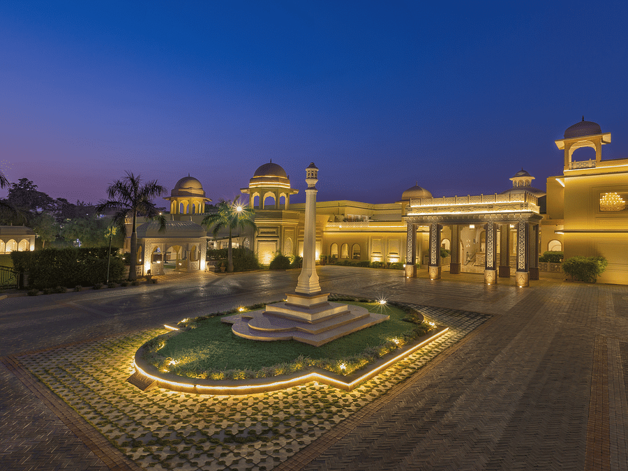 Front view of Heritage Village Resorts & Spa, Manesar with driveway, central fountain, and landscaped entrance in night.