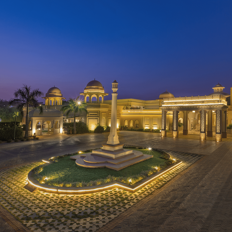 Front view of Heritage Village Resorts & Spa, Manesar with driveway, central fountain, and landscaped entrance in night.