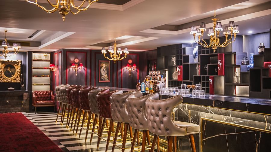  Luxurious bar at The Manor Sports & Wellness Hotel with tufted bar stools, golden chandeliers, and a black marble counter set against dramatic, opulent interiors.