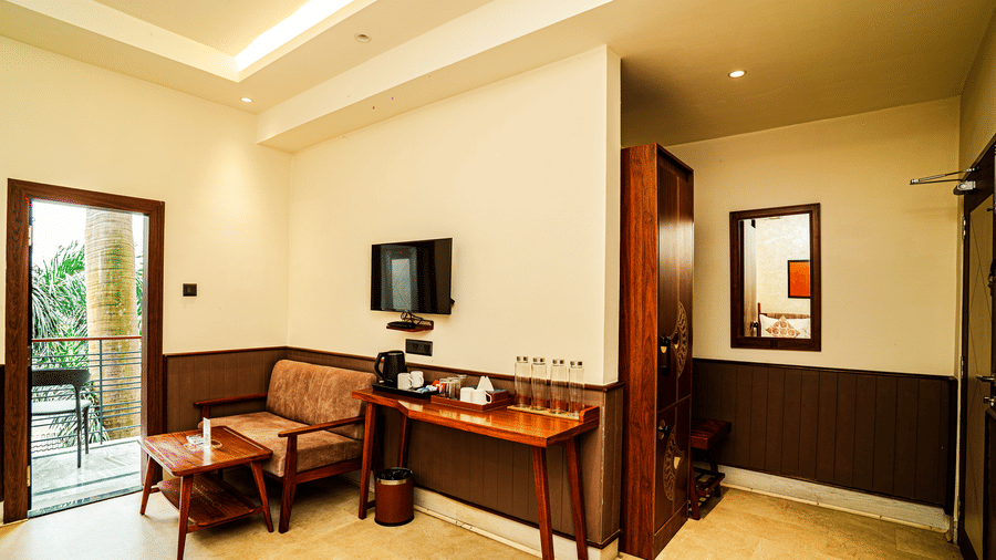 The seating area in our Deluxe Four Bedded Room featuring a cushioned chair, a coffee table, a wall-mounted television, and a balcony overlooking the hotel | Hotel Sonar Bangla, Tarapith