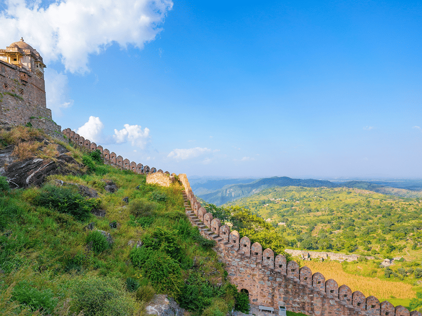 Aerial view of Kumbhalgarh Fort walls winding across green hills, with distant valleys beneath a bright blue sky and scattered clouds.