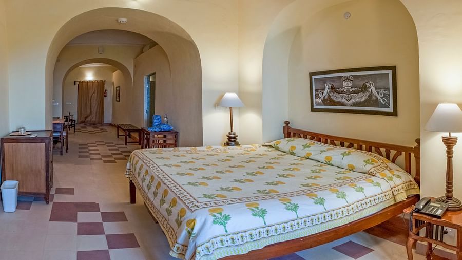 Hotel room with a bed featuring floral bedding, bedside tables with lamps, a desk, artwork, and arched doorways leading to a hallway at Tijara Fort-Palace, Alwar.
