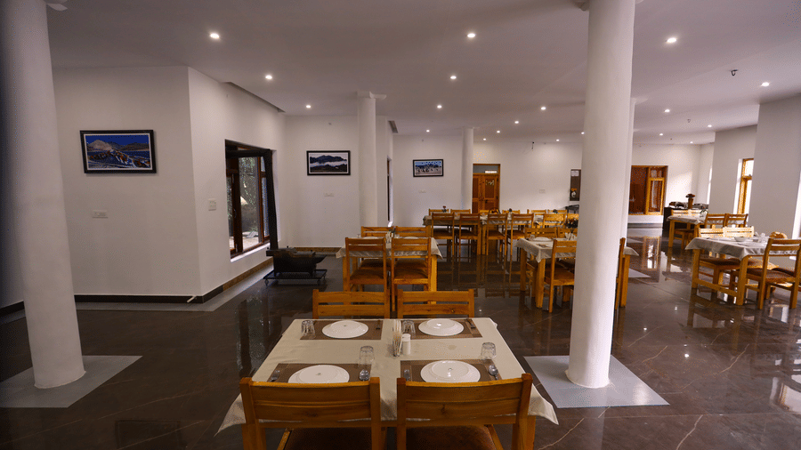 A dining space with chairs, plates placed on tables, a person serving in a cup with saucer, and wall arts at Indus Serinity, Leh Ladakh.
