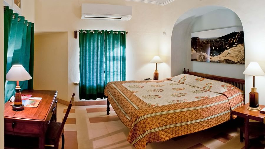 Room with bed, patterned bedding, curtains, furniture including desk and tables, lamps, wall-mounted air conditioner, and artwork in arched alcove - Tijara Fort-Palace - 19th Century, Alwar.