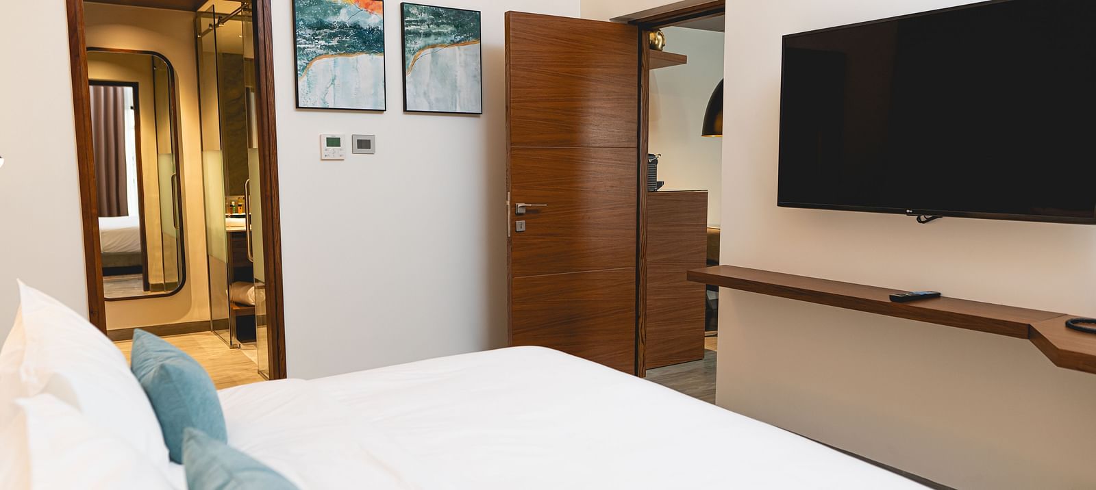 KertelSuites room with a neatly made bed, pillows, a wall-mounted flat-screen TV, wooden door, desk, and stylish decor. The bathroom is visible through an open door in the background