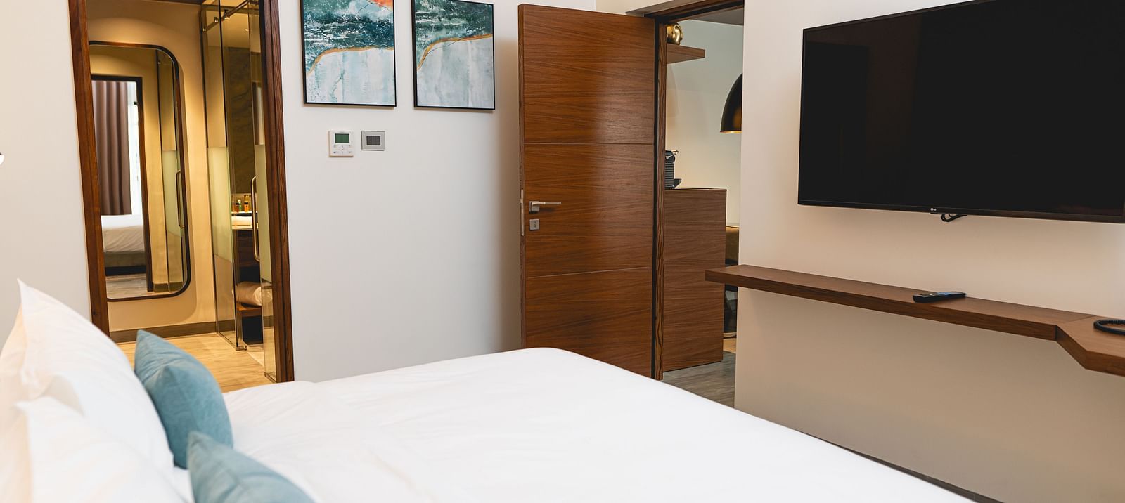 KertelSuites room with a neatly made bed, pillows, a wall-mounted flat-screen TV, wooden door, desk, and stylish decor. The bathroom is visible through an open door in the background