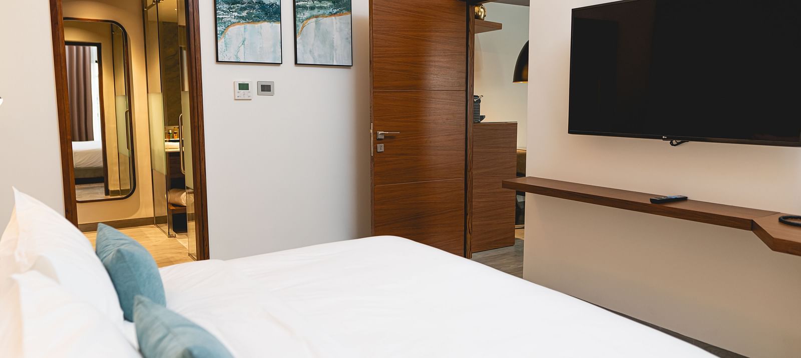KertelSuites room with a neatly made bed, pillows, a wall-mounted flat-screen TV, wooden door, desk, and stylish decor. The bathroom is visible through an open door in the background