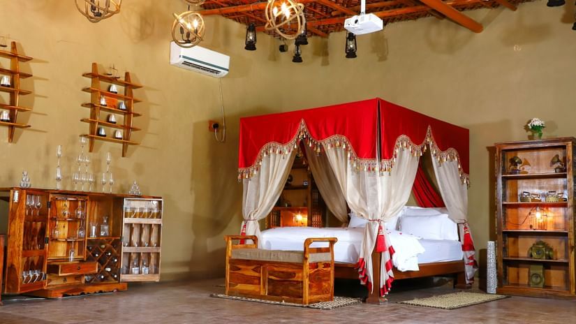 Spacious rustic room with red-canopied four-poster bed, wooden furniture, decorative shelves, warm lighting, and traditional ethnic-inspired décor at Themis Mudhouse- An Ethnic Resort & Cultural Dining